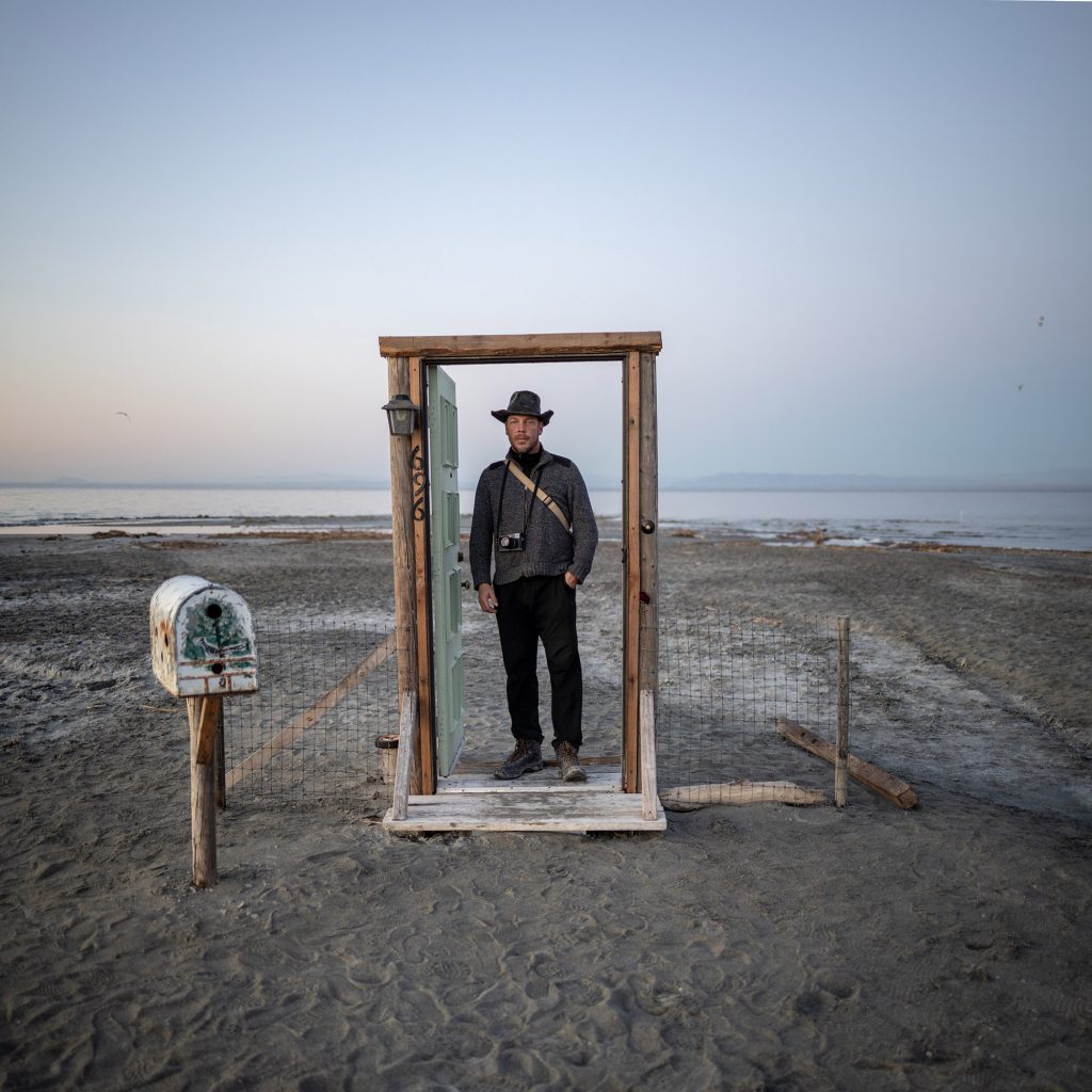 Uwe H. Martin standing in a freestanding doorframe on the shore of the Salton Sea at dusk, camera around his neck. Bombay Beach, California. Photo: Dennis Dimick.